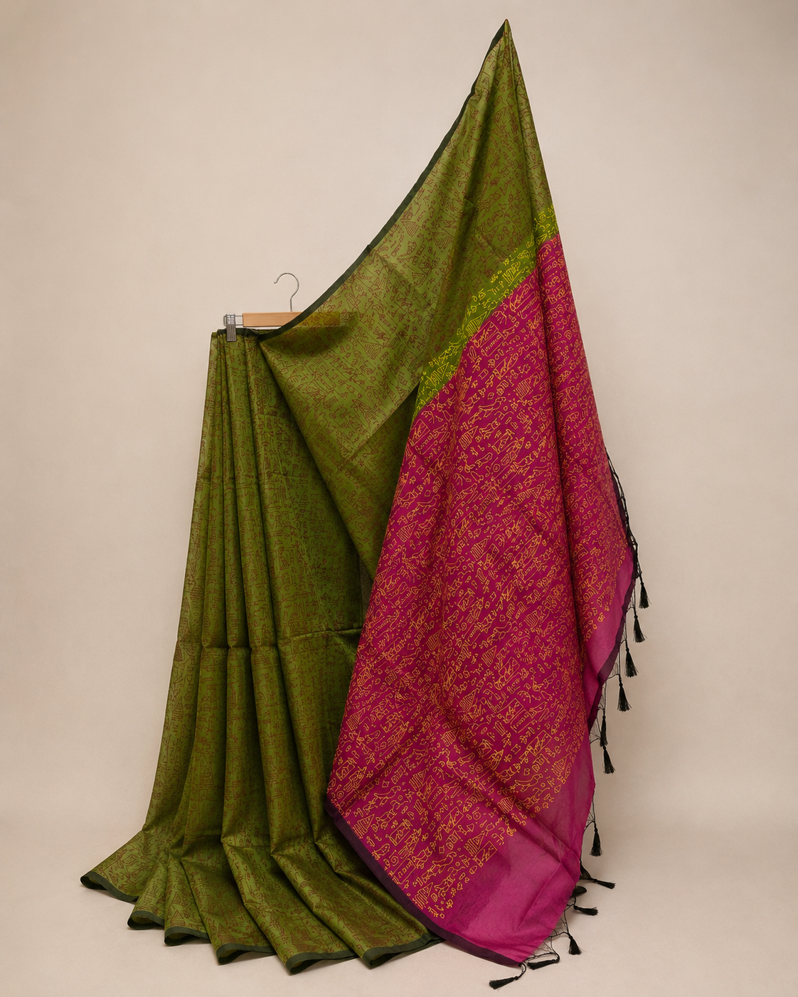 An olive green cotton saree draped elegantly on a hanger against a neutral beige background, featuring a vibrant magenta-pink border with gold floral patterns and black tassels.