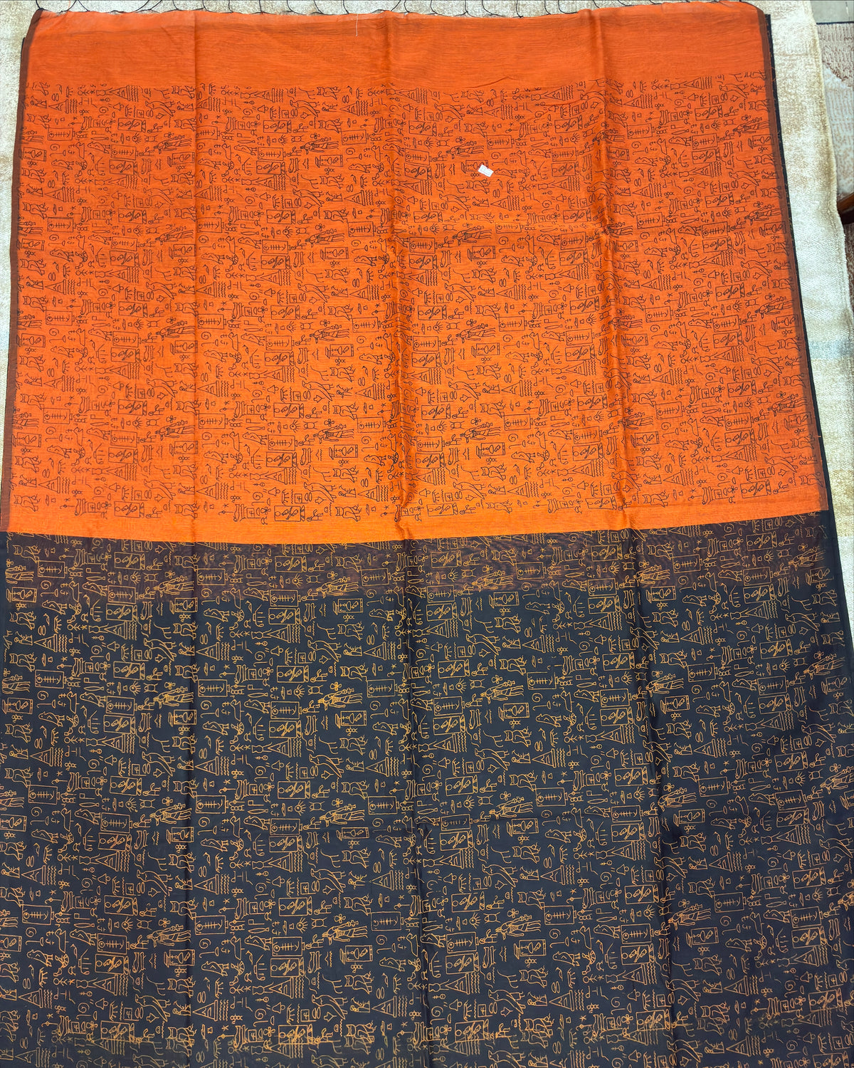 A rectangular orange and black printed fabric, likely a saree or scarf, laid flat on a stone surface. The top half is bright orange with dense black text-like print, and the bottom half is black with a gold-toned pattern.