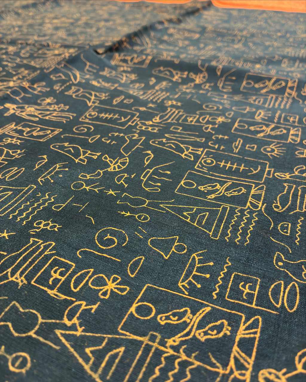 A close-up of dark teal fabric with an all-over gold embroidered pattern of abstract symbols, characters, and small illustrated motifs.
