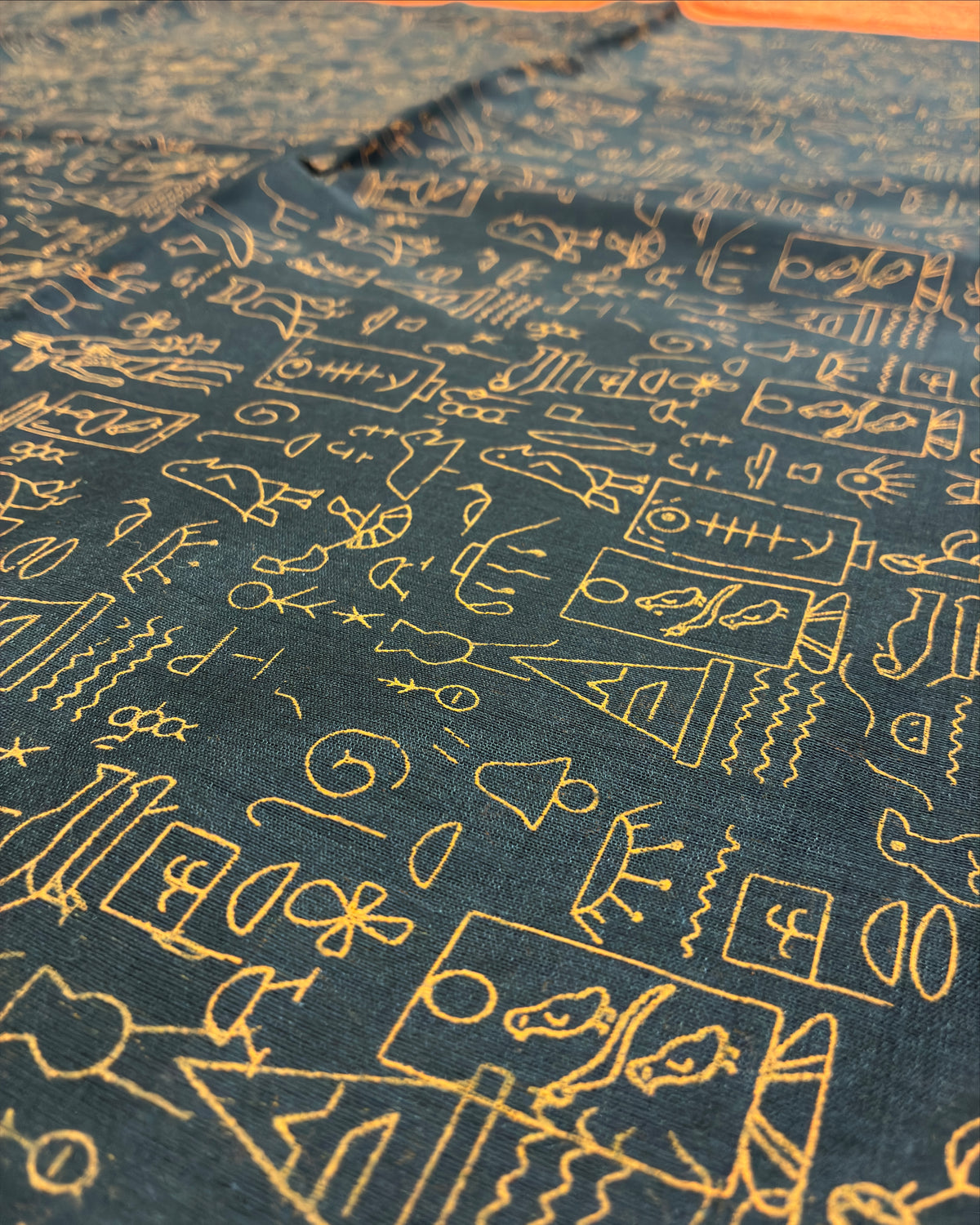 A close-up of dark teal fabric with an all-over gold embroidered pattern of abstract symbols, characters, and small illustrated motifs.