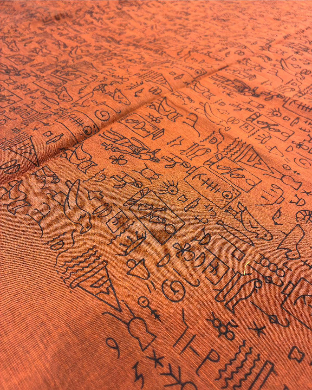 Close-up of a textured orange fabric printed with dense black line-art symbols, characters, and abstract markings across the surface.