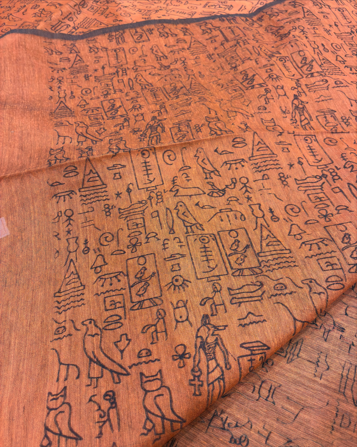 A close-up of a peach-orange fabric printed with black ancient Egyptian hieroglyphs and symbols, showing a draped fold.