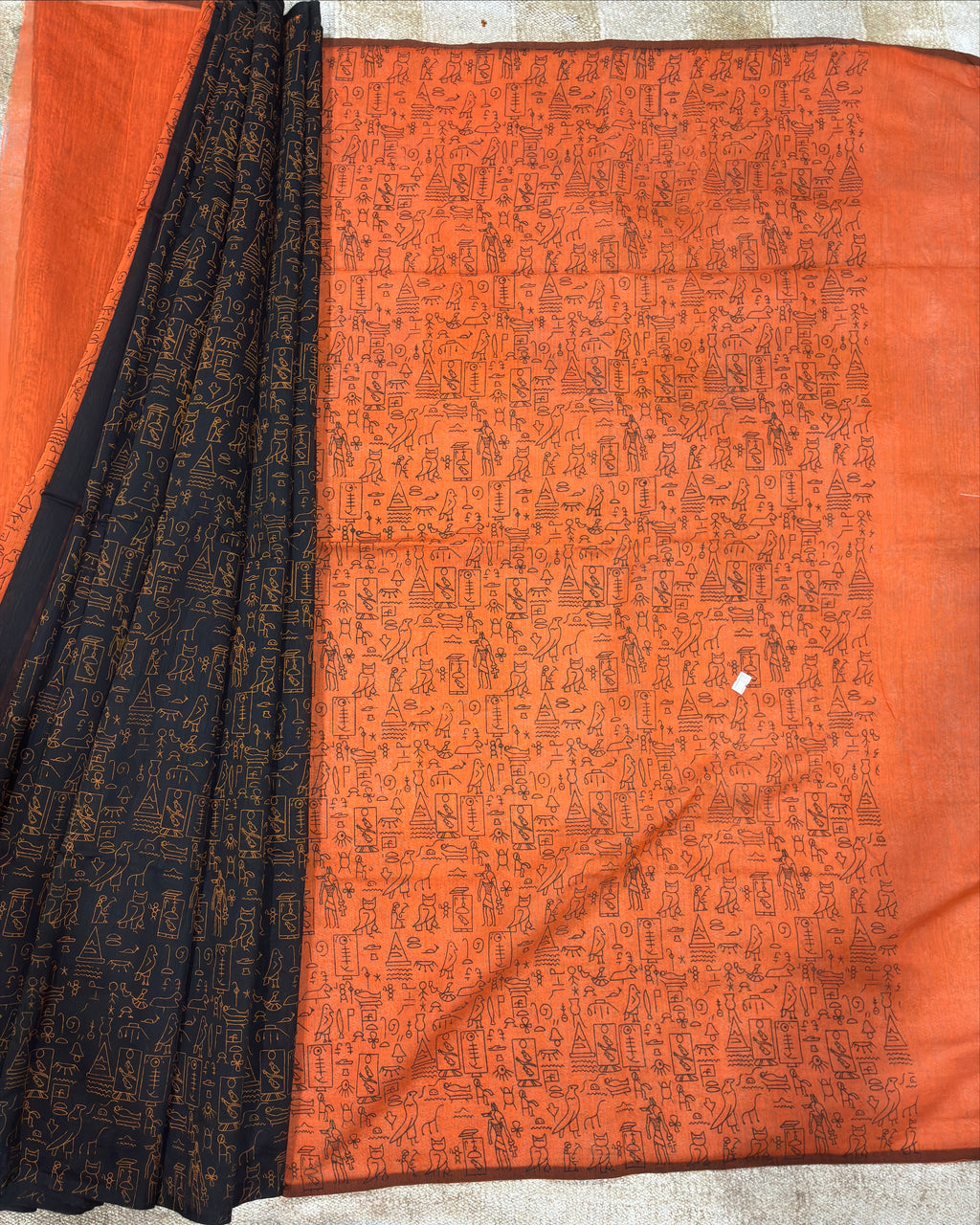 An orange and black printed fabric, likely a saree or textile, laid flat on a light-colored floor. The main body is a vibrant orange with dense black traditional patterns, and a black border with gold-toned motifs is draped on the left side.