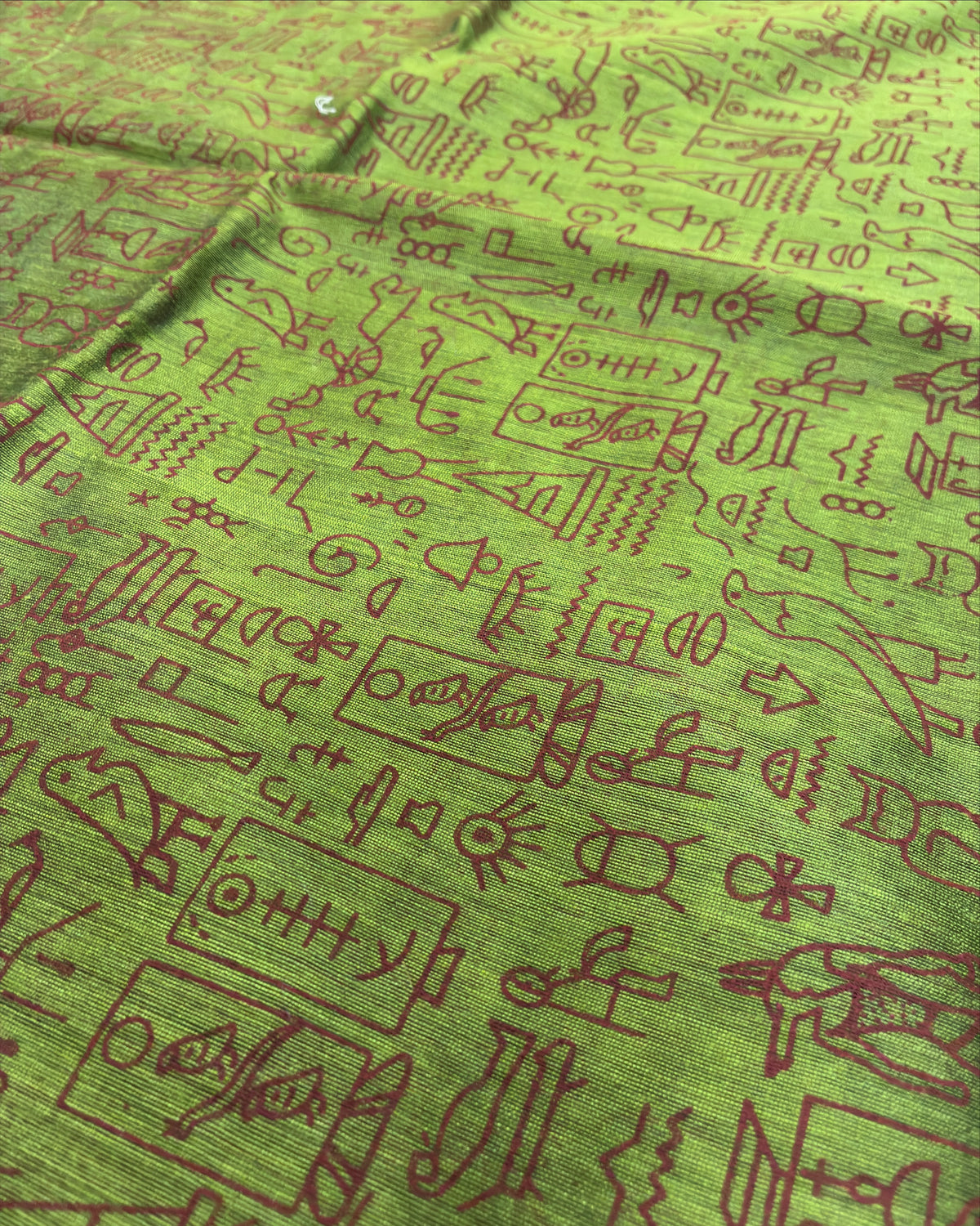 A close-up of a green fabric with an all-over brown line-art pattern of ancient script, symbols, and illustrations, featuring a small silver button near the top edge.