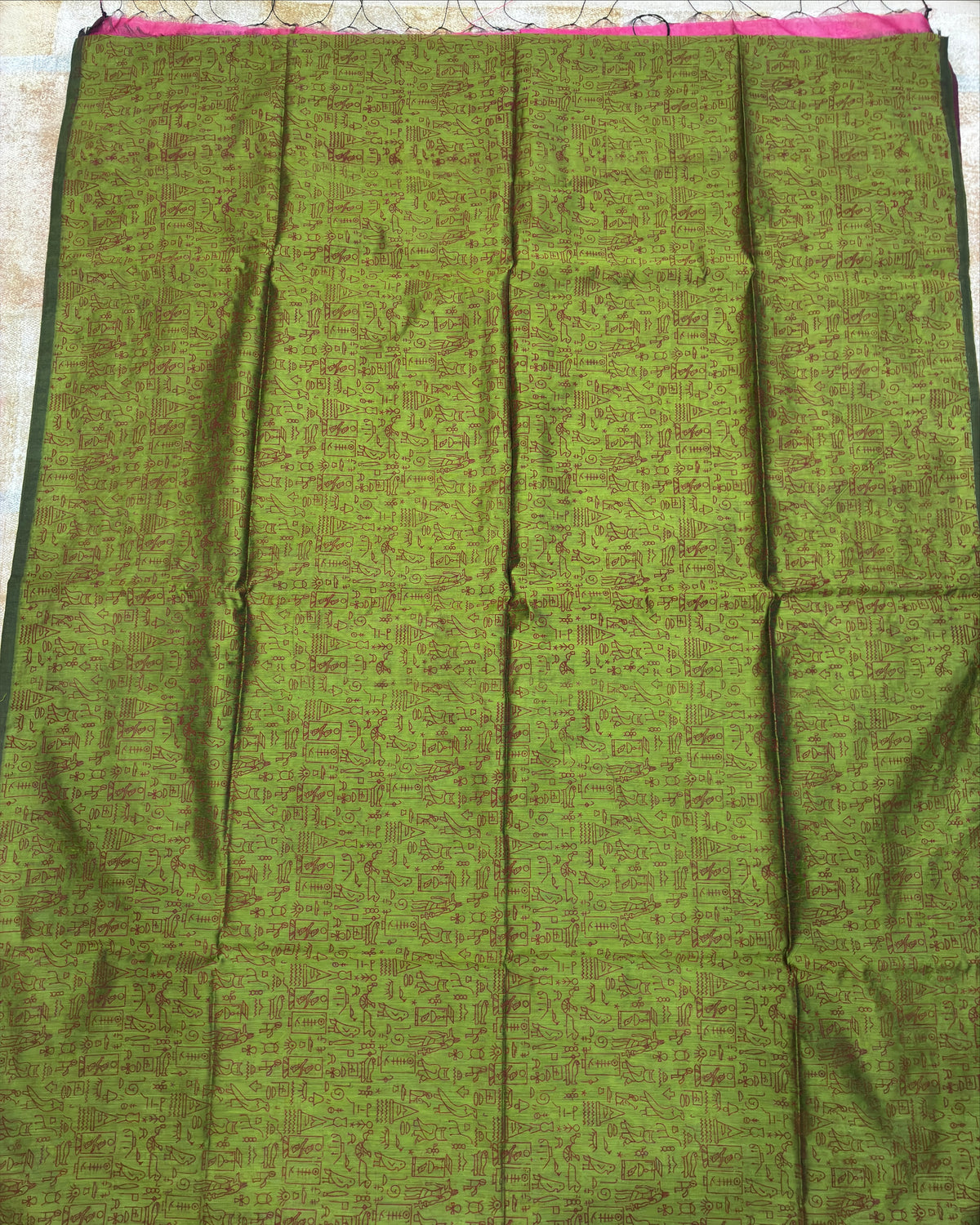 An olive green cotton saree fabric with a subtle brown floral pattern, laid flat on a light-colored tiled floor. The fabric is folded in a grid pattern, showing its texture and color, with a small glimpse of a pink border at the top edge.
