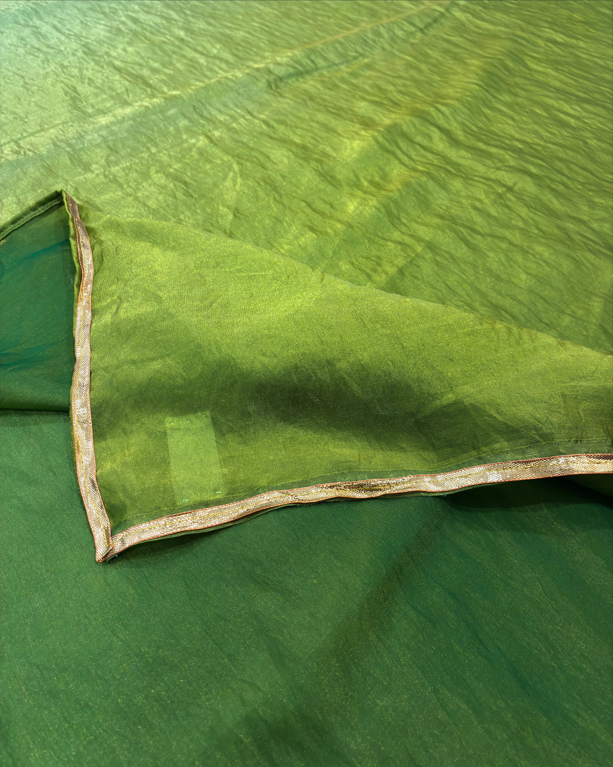 A close-up of a green fabric, likely a saree or textile, with a folded corner revealing a metallic gold border.