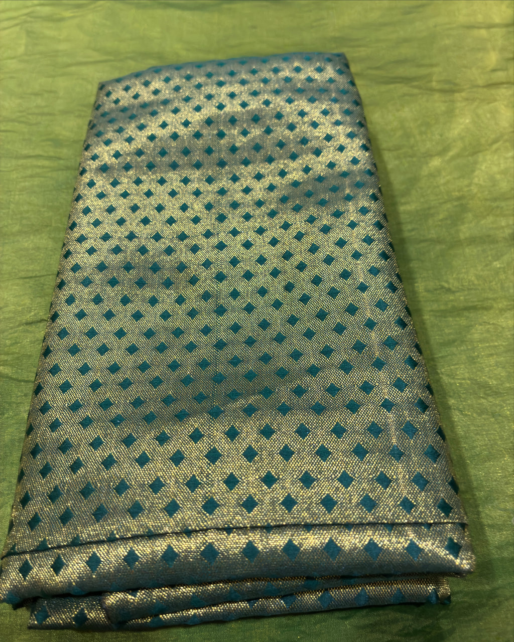 A folded piece of metallic silver fabric with a repeating pattern of small teal blue diamonds, displayed on a textured olive green background.